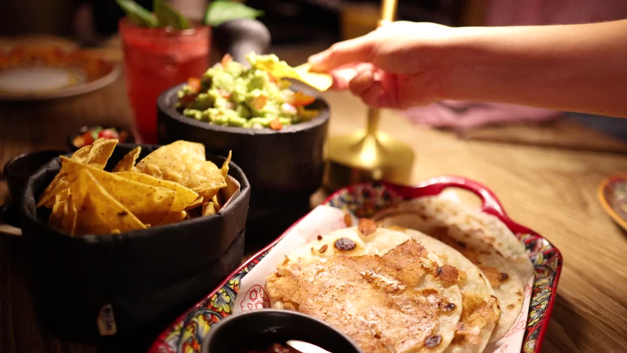 A hand dips chips into guacamole at a warmly lit table with Mexican dishes in a Bangkok restaurant