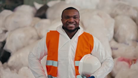 Portrait-of-a-Happy-man-with-Black-skin-in-a-white-protective-uniform-who-holds-in-his-hands-a-protective-white-helmet-and-an-orange-vest-on-it.-He-stands-near-large-bags-of-cellophane-and-polyethylene-at-a-waste-recycling-plant