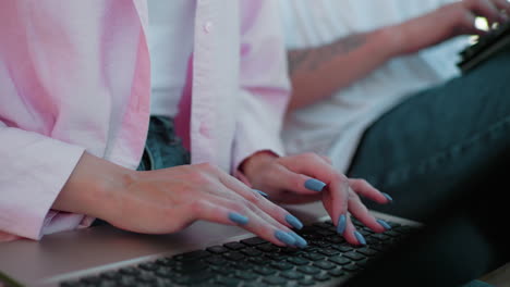 close-up of woman in pink top with polished nails and jeans typing on laptop while showing something on tablet to colleague seated next to her who types quickly