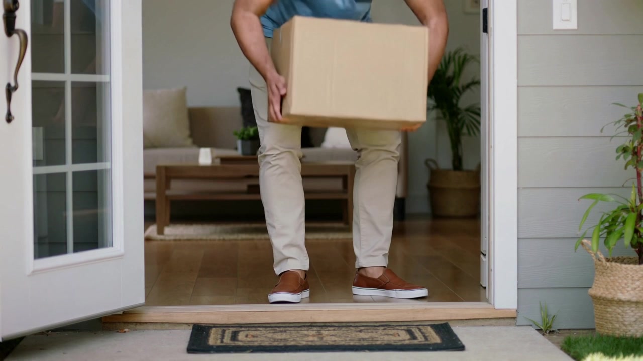 Picking up man in blue tee, tan chinos carrying cardboard box over threshold into home