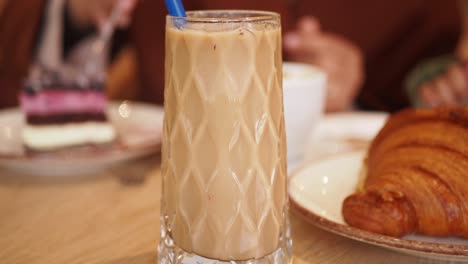 a glass of iced coffee on a table with a croissant and a piece of cake