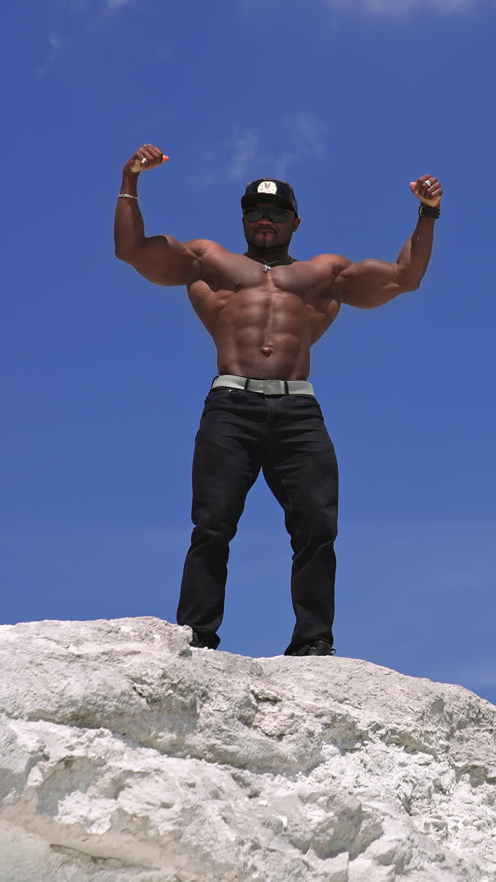 African american sportsman posing on blue sky background. Dark skinned athlete showing his strong arm muscles outdoors. View from below. Vertical video
