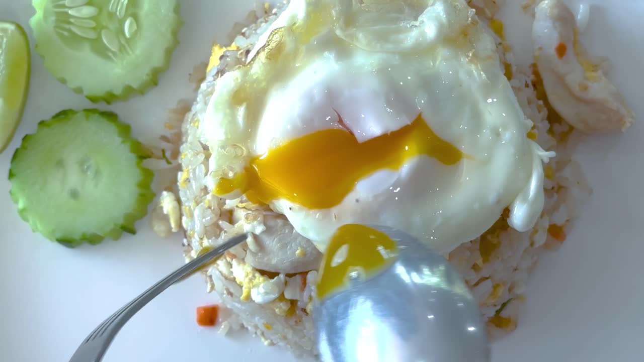 A spoon and fork break a runny egg yolk over fried rice, accompanied by sliced cucumbers.