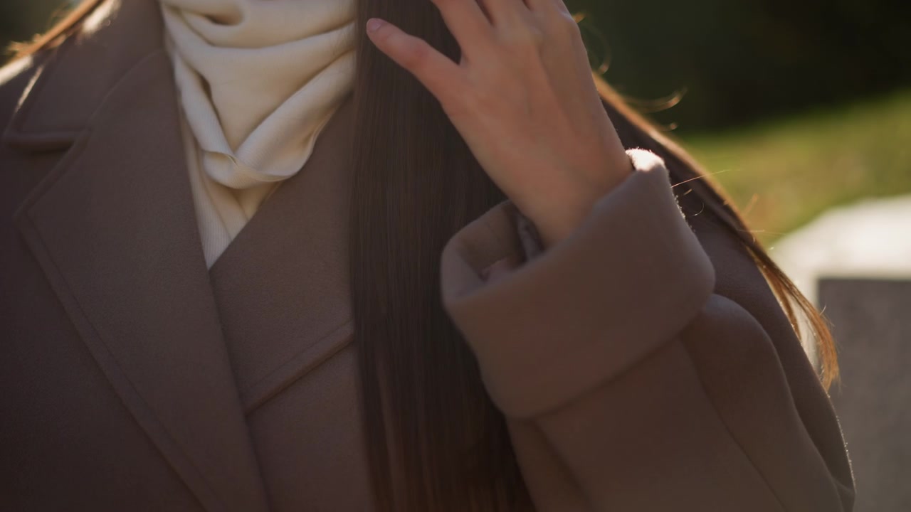 Primo piano di una donna che si sistema i capelli mentre indossa un cappotto marrone e una sciarpa color crema in una luce naturale soffusa. Il movimento della mano è lento e deliberato, concentrandosi sull'interazione tra le dita e i lunghi capelli.