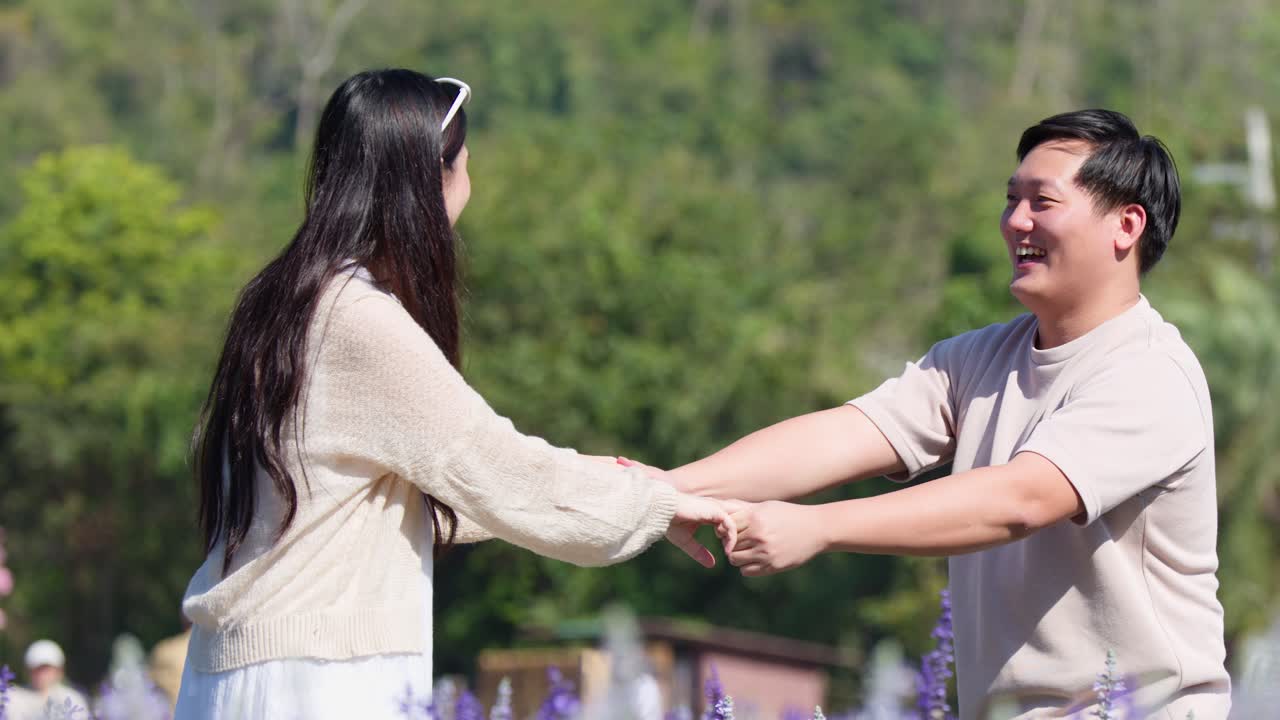 คู่รักยิ้มแย้มจับมือกันเล่นในสวนดอกไม้สีม่วงที่มีแดดส่องถึง ถ่ายภาพระยะกลางแบบถือด้วยมือ