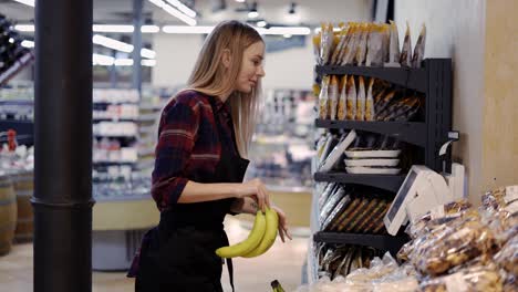 Trabajadora-De-Tienda-En-Delantal-En-El-Departamento-De-Frutas-Del-Supermercado-Que-Pesa-Plátanos