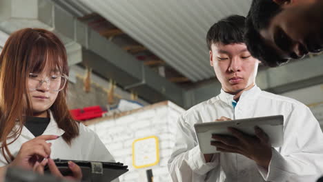 inventor in lab coat takes notes on tablet while working in mechanical workshop, partial view of another person nearby, industrial workspace filled with engineering components in background