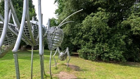 camera moves smoothly around large metallic insect sculptures on grassy lawn, surrounded by lush trees in bright natural daylight at durham botanical gardens