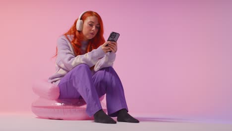 studio shot of young gen z woman wearing headphones streaming music to mobile phone 3