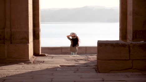 caucasian woman with hat running through the ruins of temple of philae with lake in background in aswan, egypt