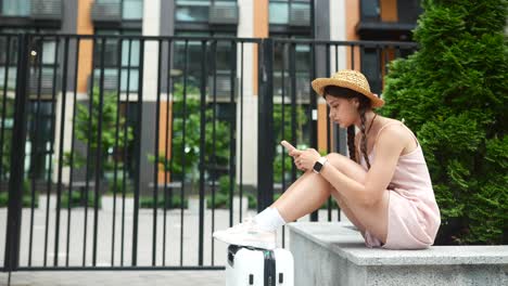 young woman sitting outdoors with suitcase and smartphone