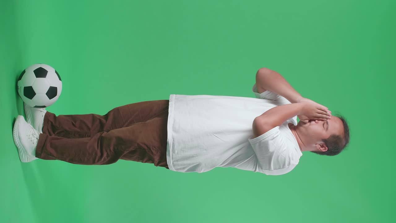 눈이 닫힌 몸, 공을 들고 있는 아시아인 남자가 손을 들고 팀을 위해 기도하면서 녹색 스크린 배경에서 축구 경기를 응원하고 있다.