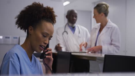 Mixed-race-hospital-worker-in-reception-talking-on-phone-with-doctors-in-backgrund