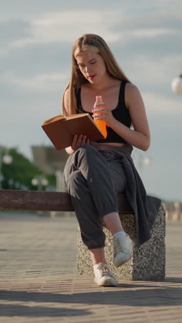 mujer sentada al aire libre con las piernas cruzadas, sosteniendo un libro en una mano mientras bebe de una botella de jugo en la otra, continuando leyendo, el fondo borroso presenta postes de lámpara y personas caminando