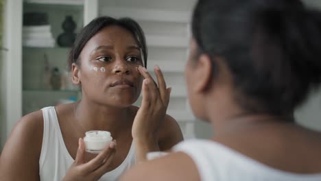 Junge-Afroamerikanische-Frau,-Die-Im-Spiegelbild-Gesichtscreme-Aufträgt