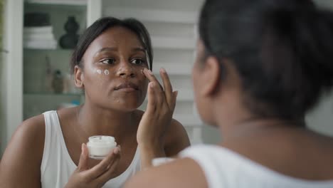 young african-american  woman applying face cream in the mirror reflection