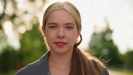 close-up of lady looking directly into camera with a soft smile, her face gently illuminated by warm natural sunlight from behind, blurred greenery in the background