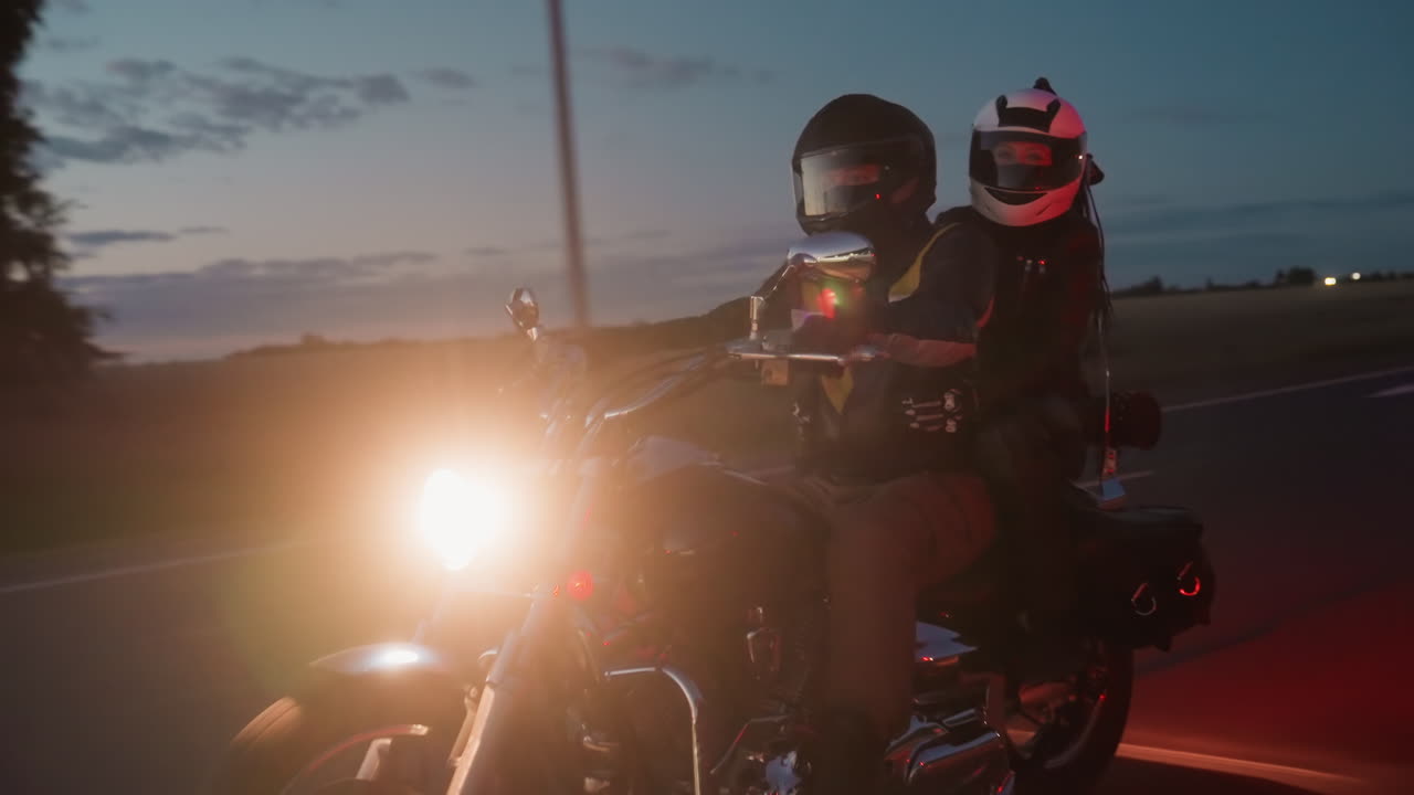 Motorcycle couple wearing helmets riding together on evening highway, bright headlight shining as they enjoy road trip at dusk, filmed from passing car with scenic countryside