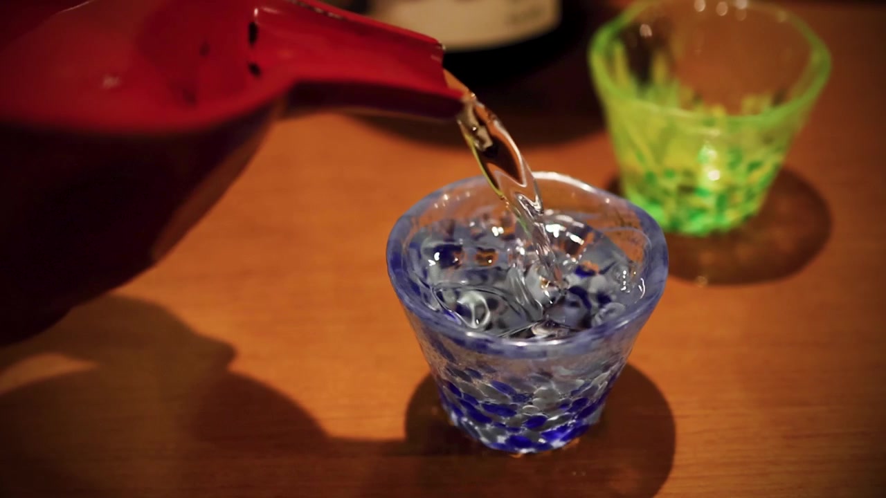Sake giapponese limpido viene versato da una brocca di ceramica rossa in una tazza di vetro blu.