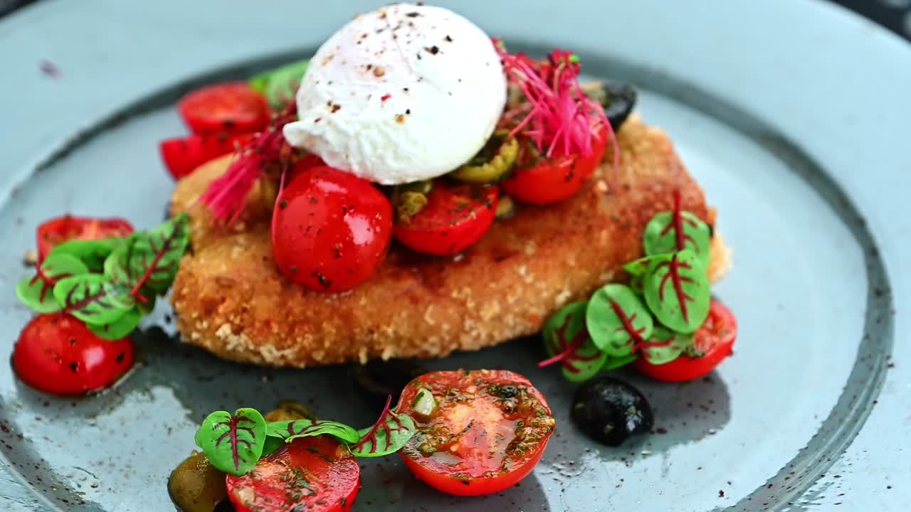 Piatto delizioso con un alimento fritto, pomodori ed erbe aromatiche su un piatto grigio. Un piatto contiene un alimento fritto con sopra un uovo in camicia, circondato da pomodorini e verdure.