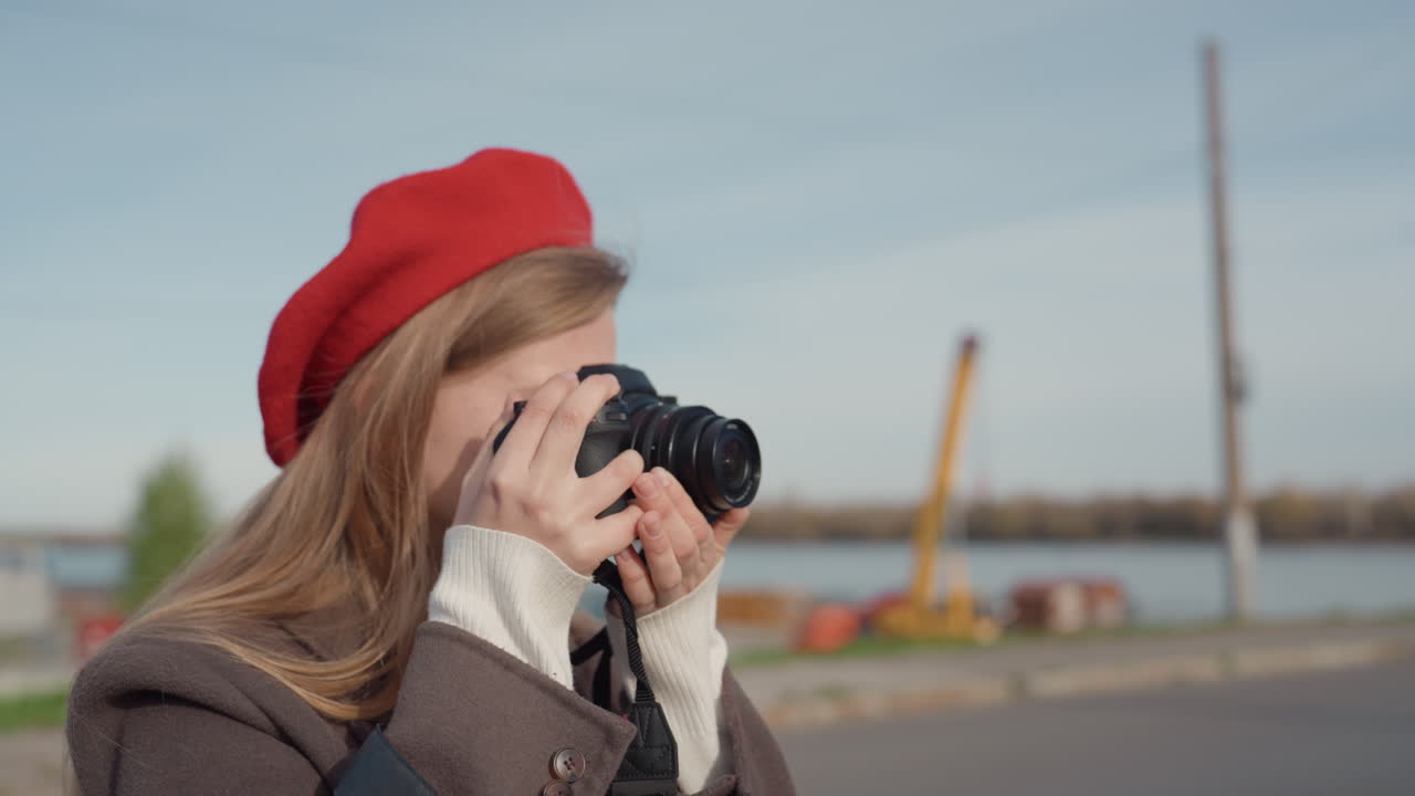 Giovane artista scatta fotografie dettagliate vicino a una zona industriale in autunno. Con la sua macchina fotografica retrò, si concentra con entusiasmo sul paesaggio lungo il fiume contro uno sfondo urbano.