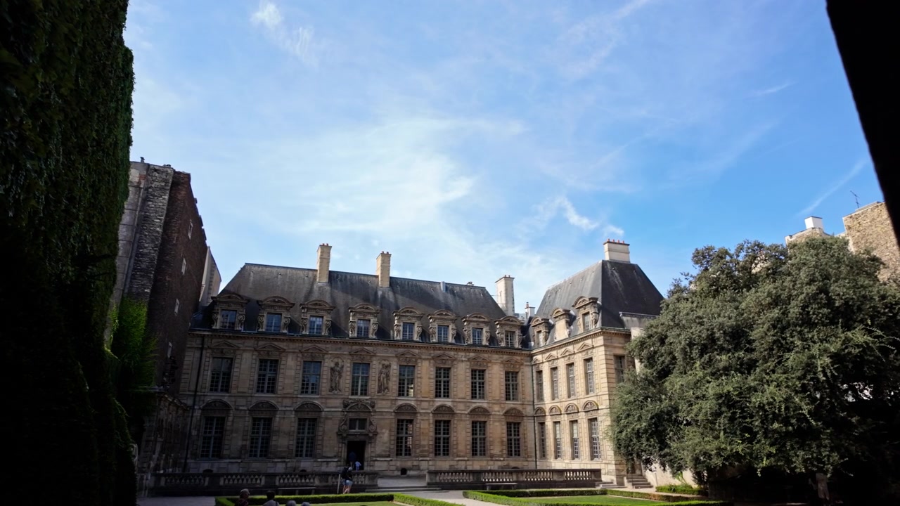 Elegante palazzo parigino e cortile con giardino formale incorniciati da un arco scuro sotto un cielo diurno di un blu brillante