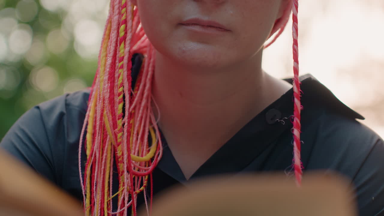 Una mujer lee pensativa, una mujer con un cabello vibrante absorta en la lectura en un parque, una persona con trenzas multicolores muestra una serena concentración mientras está absorta en un libro al aire libre