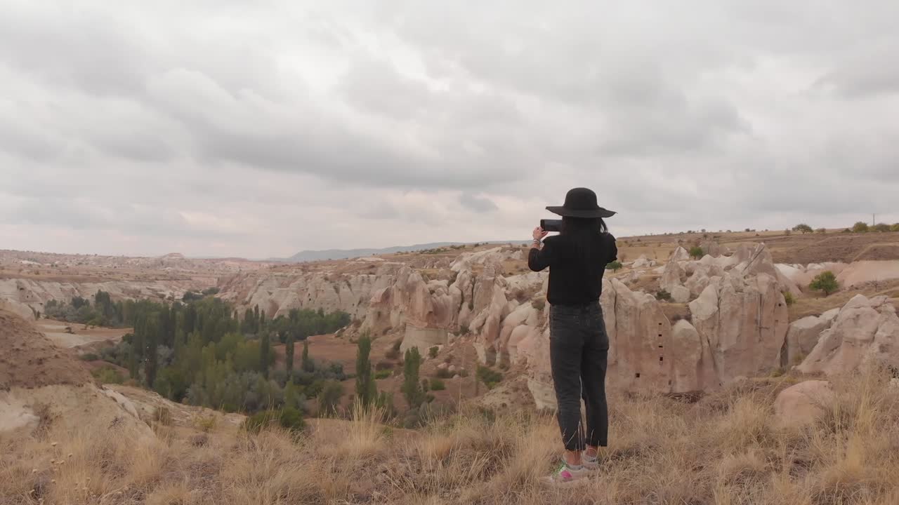 Vista com zoom de jovem turista caucasiana com roupas casuais, registrando a natureza nas férias com smartphone na mão.