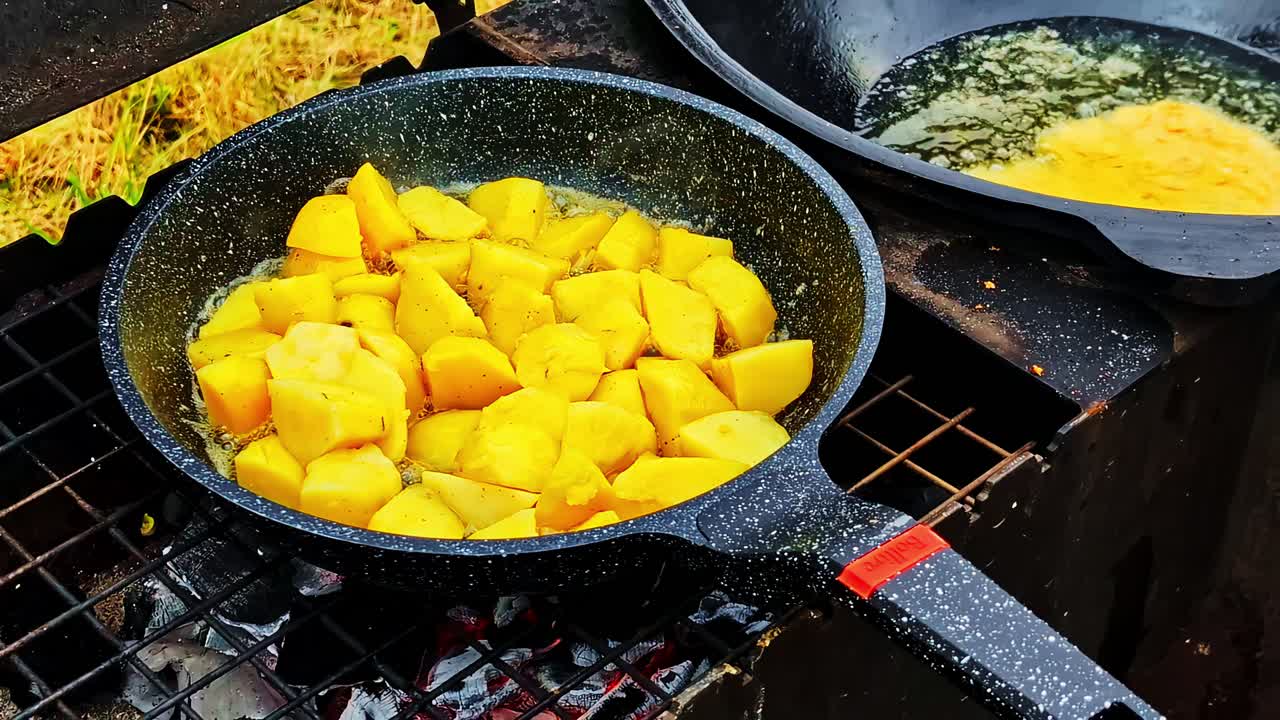 Sizzling pan of fried potatoes cooking outdoors on open charcoal flame, yellow color intense and frying food