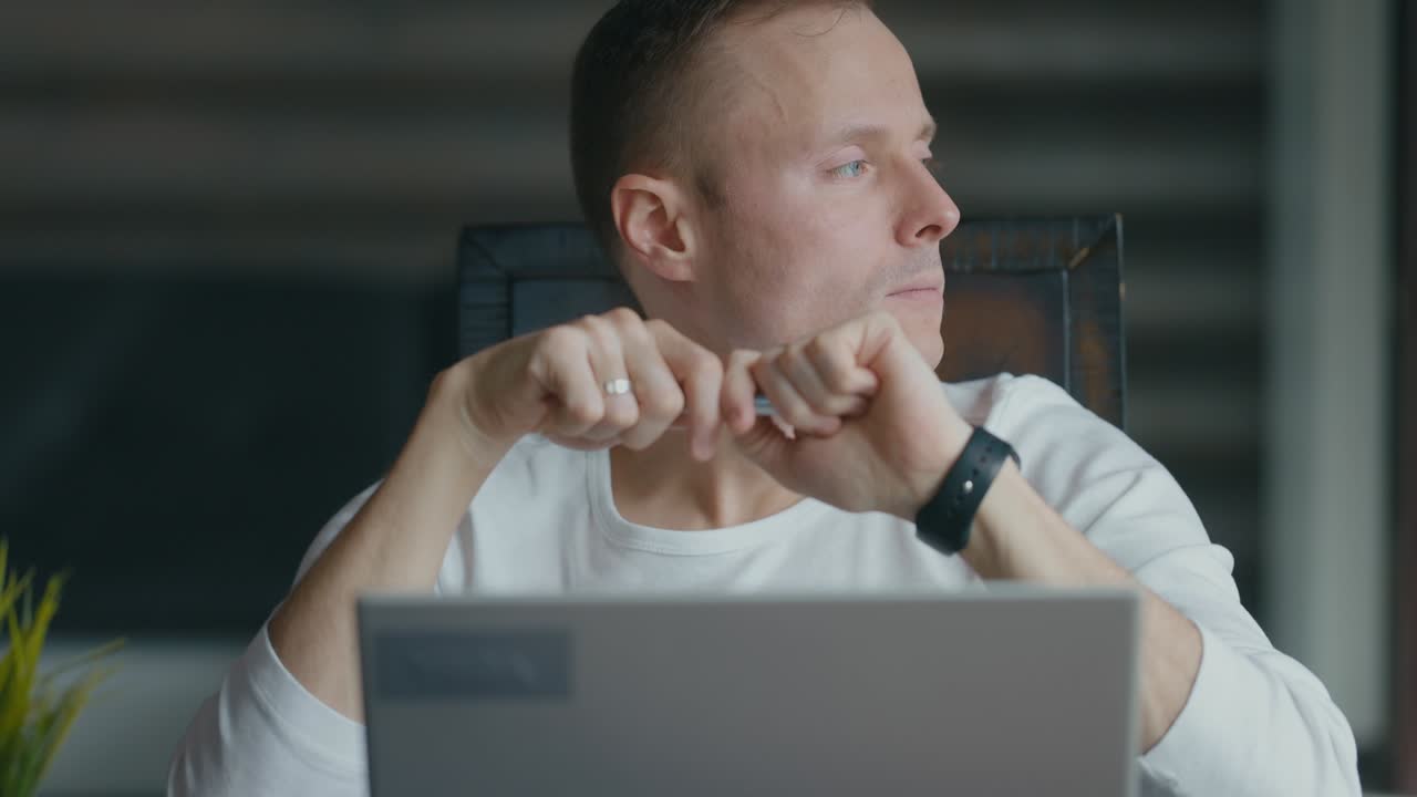 Un giovane uomo serio siede al tavolo e guarda il laptop. Gira la penna tra le mani.