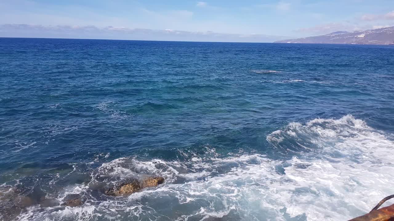 Locked shot of ocean waves crashing on rocky shore. Waves and foam in the ocean on a warm summer day. blue foam of the pacific ocean. Blue skies with white clouds.