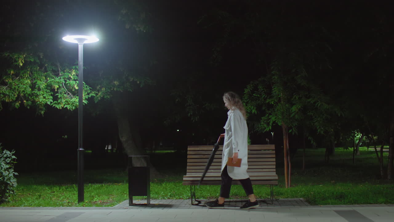 College student walks up to wooden bench under streetlight holding umbrella and book, drops umbrella on bench, sits down crossing legs, opens book to read surrounded by nighttime park greenery