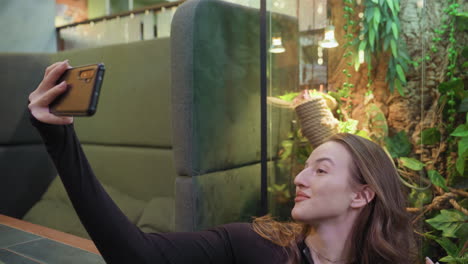 lady holding smartphone and taking selfie while seated near glass enclosure filled with lush greenery, with relaxed expression and one hand adjusting hair under warm lighting