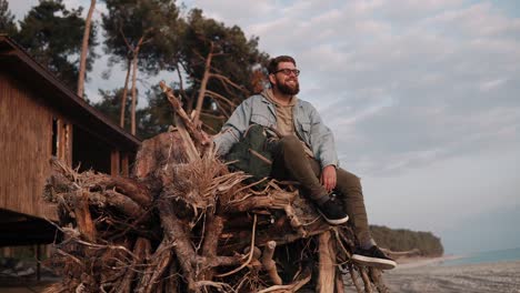 young bearded man is sitting on wooden branches on sand beach