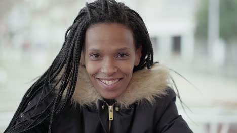 afro-american happy young girl with braids in grey jacket with fur hood sitting outside