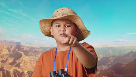 asian boy with a hat and binoculars pointing his finger towards you and furiously speaking against blaming you while traveling at the top of mountain. boy researcher, travel adventure, close up