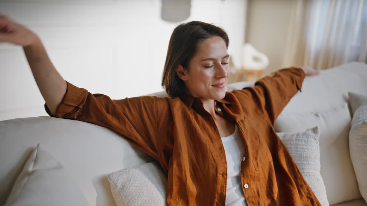 Smiling brunette relaxing sofa stretching arms on white soft pillows closeup