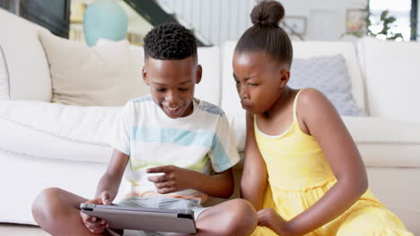 african american siblings using tablet together, sitting on floor in living room