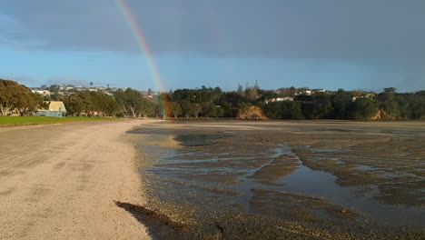 drone flying backward on a beach through rain away from a rainbow in at the end of the beach