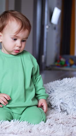 lovely calm boy of one year old sits on a soft carpet in the room. kid in light green sport suit watching videos on the phone in front of him. vertical video