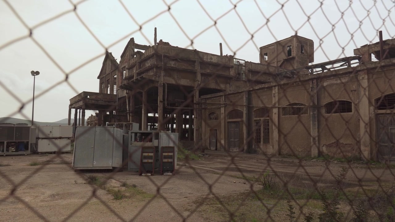 Abandoned Building Seen Through A Steel Fence At The Historic Monteponi Mining Site In Iglesias, Sardinia, Italy. Static Shot