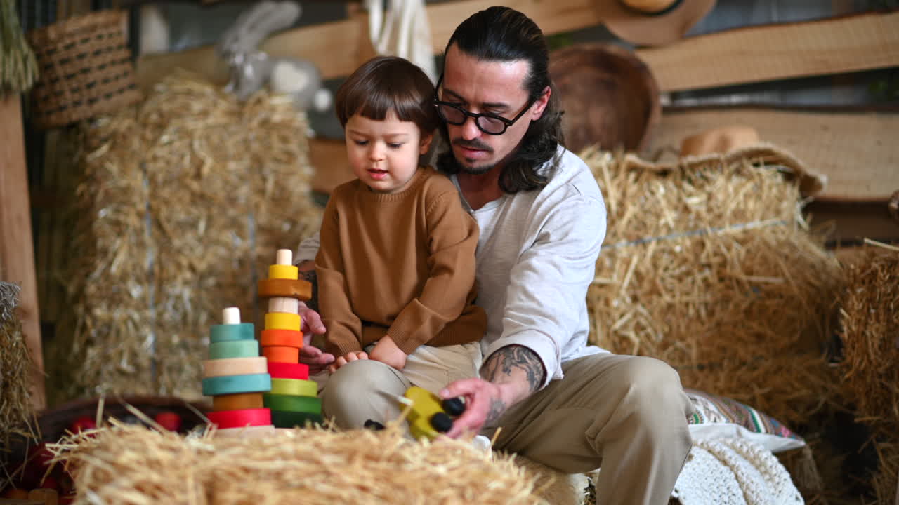 Father playing with his son with colourful, ecological wooden toys in a barn, near square hay bales