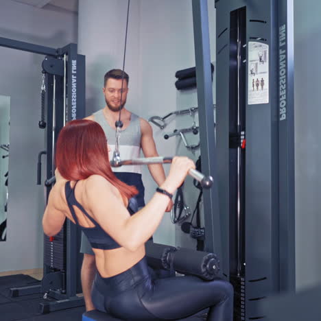 persistent woman doing her workout on a simulator. athletic female trained her muscles pulling down a bar on herself in gym. professional trainer looks at sportwoman's training indoors.