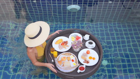 woman in mustard color bathing suit pushes floating tray of food in swimming pool