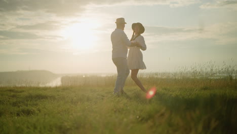 dos compañeros bailan alegremente en una colina cubierta de hierba iluminada por el sol al lado de un lago pacífico. el hombre, vestido con una camisa blanca, sombrero y vaqueros, comparte un momento tierno con su esposa, que lleva un vestido blanco