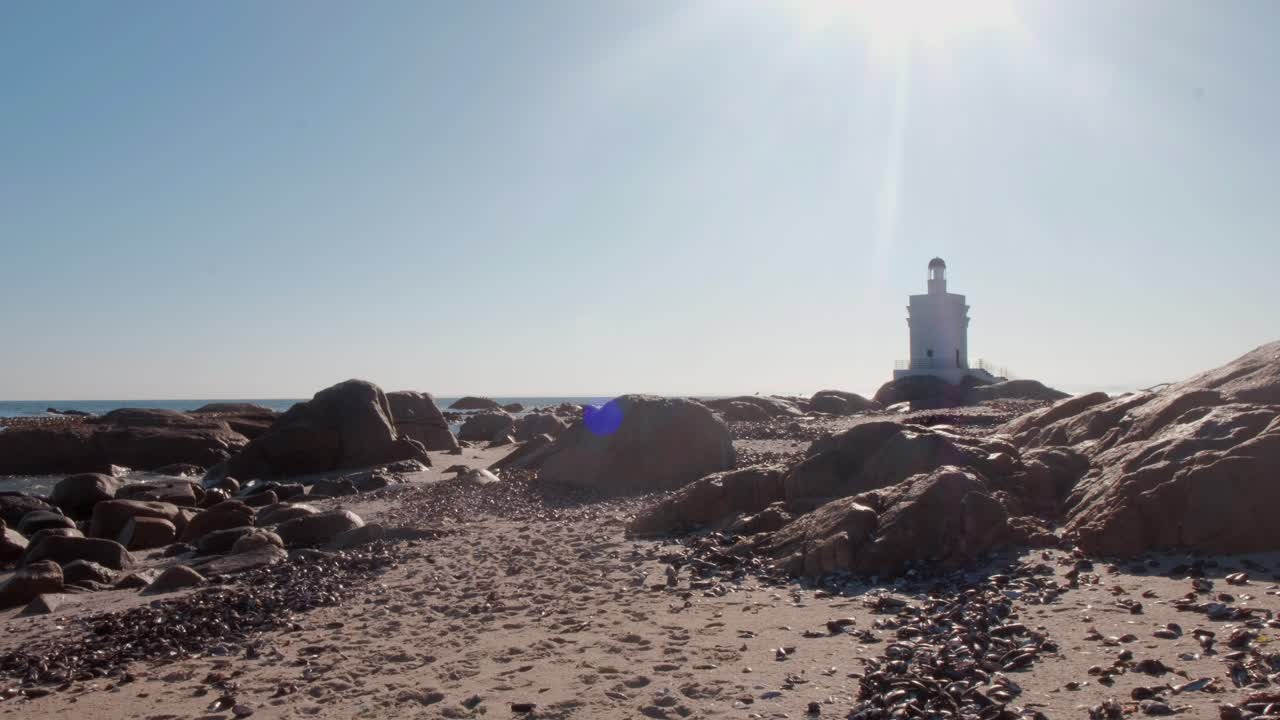 vista de un faro en la playa con olas rompiendo suavemente contra las rocas bajo el sol del mediodía