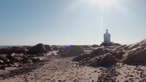 Blick-Auf-Einen-Leuchtturm-Am-Strand-Mit-Wellen,-Die-In-Der-Mittagssonne-Sanft-Gegen-Die-Felsen-Schlagen
