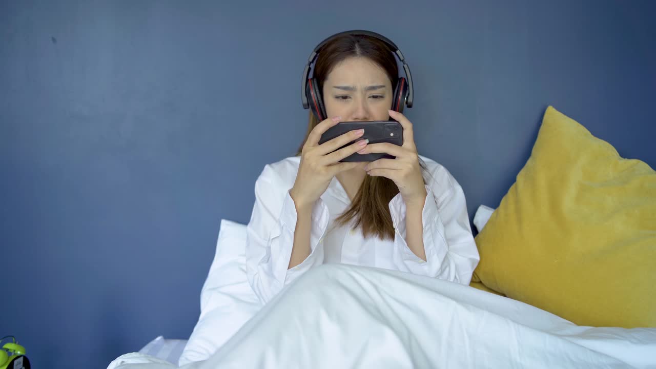 행복한 십대 여성은 헤드을 착용하고 침대에 스마트폰으로 온라인 게임을 하고 있으며, 여성은 스마트폰에서 게임을 잃었습니다.