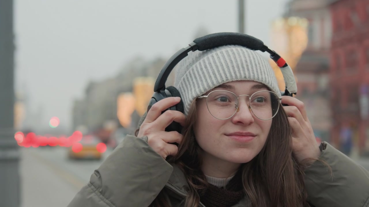 Женщина на городской улице слушает наушники. Она в серой шапке-бини и прозрачных очках, поправляет черные полноразмерные наушники холодным вечером. Размытые огни боке и движение транспорта на заднем плане. На ней парка.