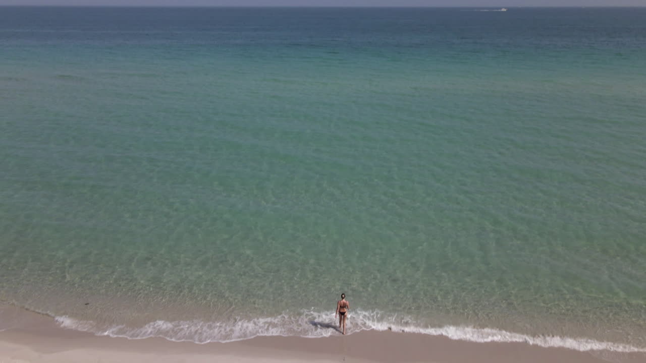 비키니 입은 젊은 백인 여성이 따뜻한 물을 향해 모래사장을 걷는다